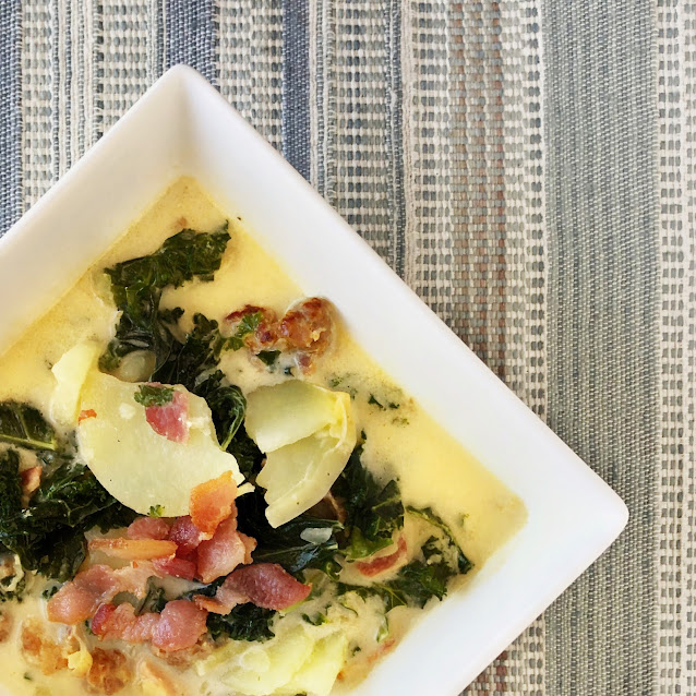creamy soup with kale, potatoes, bacon and sausage in a square white soup bowl on a blue and white stripped mat