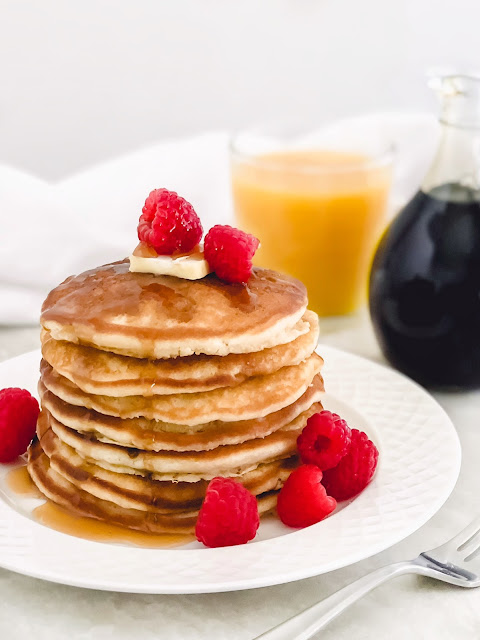 a stack of pancakes on a white plate topped with butter syrup and raspberries with orange juice and syrup in the background